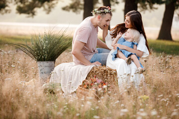 Wall Mural - Happy family together: beautiful young parents in white clothes hugging their little daughter on straw stack in the park on a sunny summer day. happy holiday. mothers, fathers, babys day