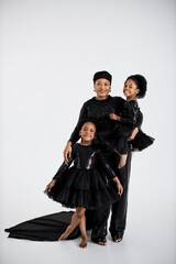 Wall Mural - Two cute african sisters standing in embrace with their charming mother over white studio background. Stunning woman with daughters wearing elegant evening clothes.