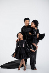 Poster - Two cute african sisters standing in embrace with their charming muslim mother in hijab over white studio background. Stunning woman with daughters wearing elegant evening clothes.