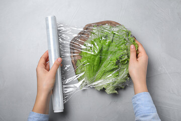 Wall Mural - Woman putting plastic food wrap over fresh lettuce at light grey table, top view