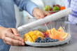 Woman putting plastic food wrap over plate of fresh fruits and berries at wooden table indoors, closeup