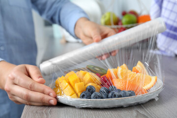 Wall Mural - Woman putting plastic food wrap over plate of fresh fruits and berries at wooden table indoors, closeup