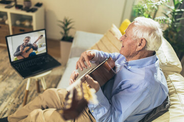 Senior man playing guitar at home using laptop for online lessons