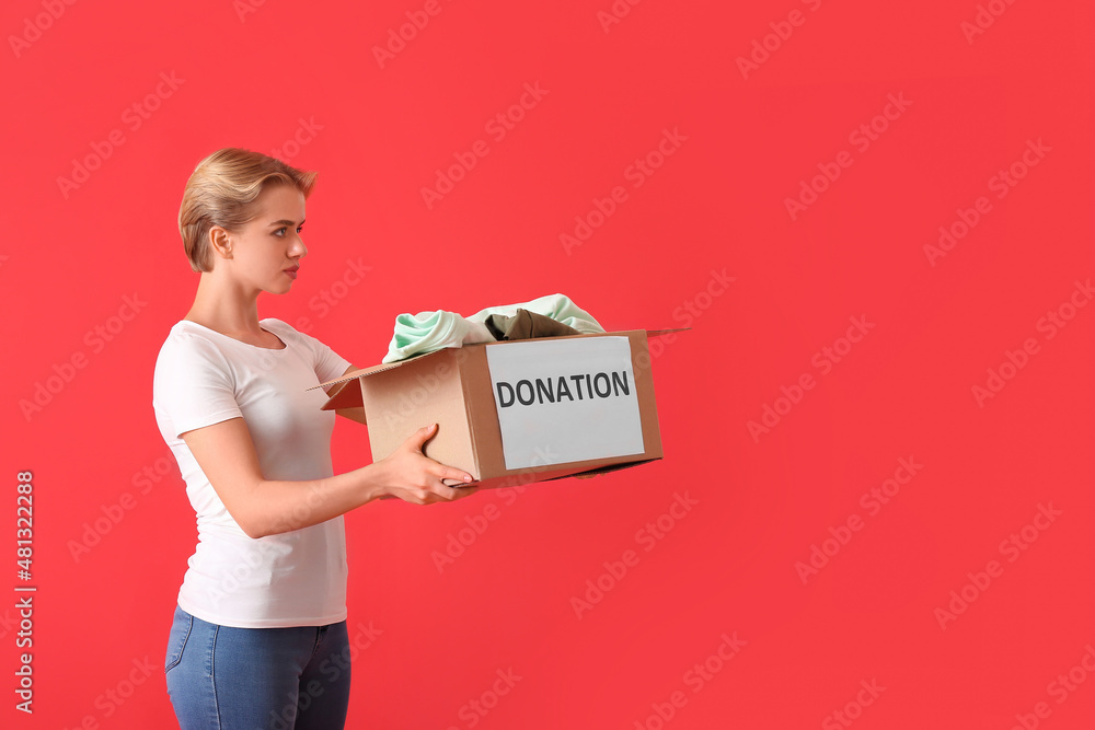 Young woman holding box with donation clothes on red background