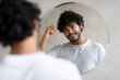 © Prostock-studio - Male haircare. Positive indian bearded guy brushing hair with hairbrush, looking at his reflection in mirror in bathroom