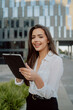 © ABCreative - pretty woman in white shirt stands in front of glass modern business building of insurance corporation company, women is holding tablet in hand, browsing web, answering email, reading messages