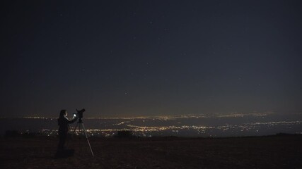 Wall Mural - Woman filming stars with a camera and a tripod at night. City lights in the background. 