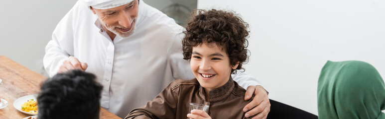 Wall Mural - mature muslim man hugging arabian grandson during dinner with family, banner.