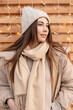 Happy fashionable girl model in trendy winter clothes with knitted sweater, vintage hat, down jacket with scarf on a wooden background with lights