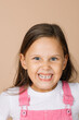 Portrait of female kid with scary, frightening grinning forced smile with all teeth with playful shining eyes looking at camera wearing bright pink jumpsuit and white t-shirt on beige background.