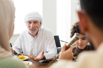 Poster - smiling muslim man pointing with hand during dinner with arabian grandson and blurred family.