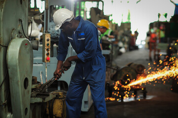 A african black man in a mechanic's suit is using a wrench to repair a machine and his back has sparks from cutting metal with a hacksaw. which is not safe from working in industrial plants.