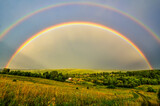 Fototapeta Tęcza - Full arc of double rainbow over summer evening rural landscape - panoramic view of hills with deep forests covered, golden colored meadow with sun beams illuminated, village near forest