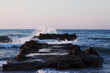 Fale morskie rozbijające się o zachodzie słońca o falochron ze skał i kamieni na Krecie w Grecji,  Agia Marina