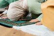 © LIGHTFIELD STUDIOS - Muslim woman praying and bending on rug near family at home.
