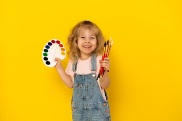 Blonde curly girl on a yellow background smiles, with paints and tassels in her hands, dressed in a denim jumpsuit and pink T-shirt