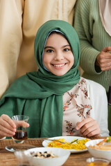 Canvas Print - happy muslim girl in hijab looking at camera while holding glass of tea near parents.