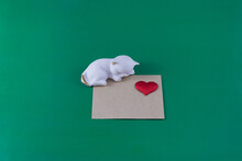 A Porcelain Figurine In The Form Of A Sleeping Kitten, A Paper Envelope And A Red Heart On A Green Background