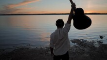 Man standing at lake waterfront and raising guitar up in the air at glorious golden sunset, Torrevieja Pink lake in Alicante, Spain - back view gimbal crane shot