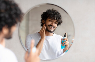 Wall Mural - Handsome indian man applying moisturizing lotion and touching beard, smoothing after shaving in bathroom