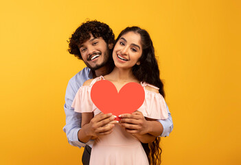 romantic indian couple holding red paper heart card, loving guy embracing his pretty woman from back