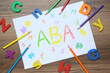 Sheet of paper with abbreviation ABA (Applied behavior analysis), plastic letters and pencils on wooden table, flat lay
