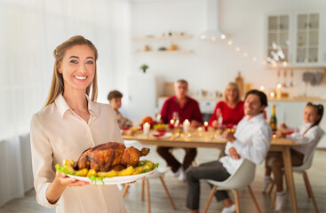 Happy young woman holding roasted turkey for Thanksgiving or Christmas, celebrating holiday with nears at home