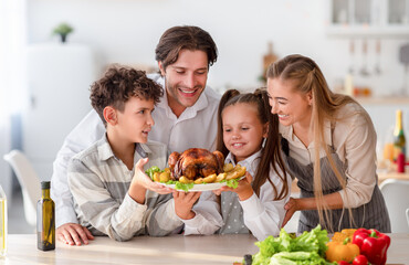 Cheerful children with parents cooking traditional dinner together, holding roast turkey for holiday meal at kitchen