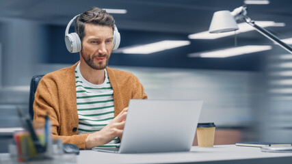 Poster - Modern Office: Handsome Businessman Sitting at His Desk Working on a Laptop Computer. Man Wearing Headphones, Listens to Music, Podcast. Motion Blur Background Showing Active Work Day.