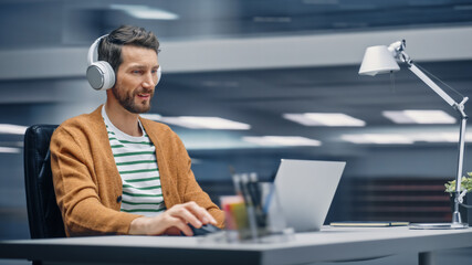 Poster - Modern Office: Handsome Businessman Sitting at His Desk Working on a Laptop Computer. Man Wearing Headphones, Listens to Music, Podcast. Motion Blur Background Showing Active Work Day.