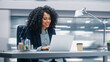 © Gorodenkoff - Modern Office: Black Businesswoman Sitting at Her Desk Working on a Laptop Computer. Smiling Successful African American Woman working with Big Data e-Commerce. Motion Blur Background