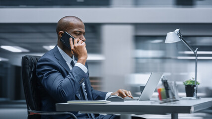 Poster - Modern Office: Productive Black Businessman Sitting at His Desk Working on a Computer, Answering Phone Call. African American Entrepreneur Wearing Suit working with Investment. Motion Blur Background