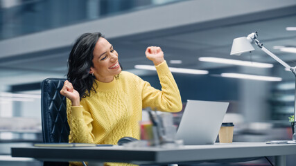 Poster - Modern Office: Happy Smiling Hispanic Businesswoman Sitting at Her Desk Working on a Laptop Computer Celebrates Victory. Latin Female Entrepreneur is a Happy Winner. Motion Blur Background