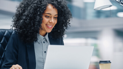 Poster - Modern Office: Black Businesswoman Sitting at Her Desk Working on a Laptop Computer. Smiling Successful African American Woman working with Big Data e-Commerce. Motion Blur Background