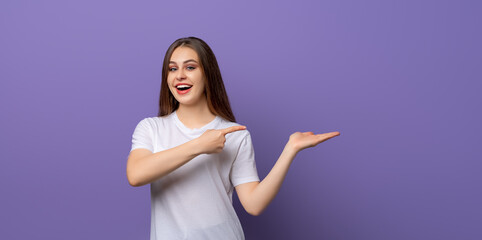 Wall Mural - Portrait of young woman surprise gazing and pointing finger aside and show something in hand over purple background