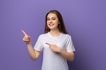 Wall Mural - Portrait of young woman pointing fingers aside and looking with positive face expression, standing over purple background