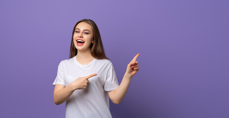 Wall Mural - Portrait of young woman pointing fingers aside and looking with positive face expression, standing over purple background