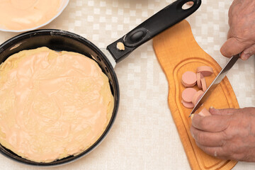 Senior woman hands cut the sausage for homemade pizza. Other ingredients and the pan with raw dough are on the table. Top view with selective focus. Process of cooking pizza, step 6