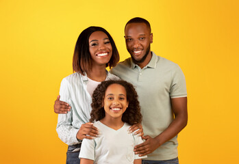 Wall Mural - Family love concept. Portrait of black family with daughter hugging and smiling at camera standing on yellow background