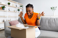 Overjoyed Afro woman unboxing carton parcel, emotional about successful shopping, satisfied with great purchase at home