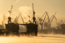 Cranes Of Baltic Shipyard In St. Petersburg In Frosty Winter Day, Steam Over The Neva River, Smooth Surface Of The River At Sunset. 