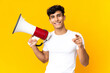 © luismolinero - Young Argentinian man isolated on yellow background holding a megaphone and smiling while pointing to the front