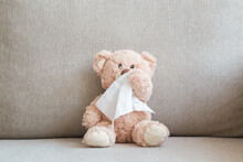 Brown Teddy Bear Sitting On Sofa And Wiping Nose With White Paper Napkin. Cold And Flu Virus. Children Healthcare Concept. Closeup. Front View.
