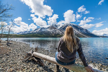 Tourist, Hiker Sitting Beside Stunning Lake In Northern Canada During Summer Time With Snow Capped Mountains In Background, Pristine Lake And Rocky Lakeshore. 