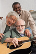 © LIGHTFIELD STUDIOS - Cheerful interracial senior friends playing acoustic guitar and hugging at home.