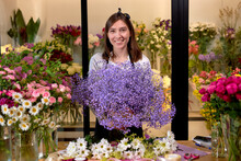 Young Woman Florist Standing In Small Flowers Shop Posing, Holding Many Prefect Blooming Purple Flowers In Hands, Smiling At Camera. Portrait. Floral Handmade, Craft, Beauty, Business Concept