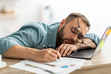 Wall Mural - Exhausted millennial man sleeping on his office desk, next to laptop and documents, tired of overworking