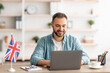 © Prostock-studio - Happy young guy with flag of Great Britain working at desk with laptop in home office