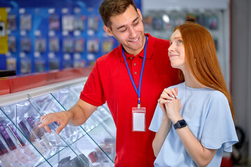 Excited Redhead Lady In Casual Wear Stand By Shelves With Smartphones In Store, With Consultant, Cute Customer Woman Is Dreaming About New Modern Mobile Phone, Consultant Help Lady To Choose