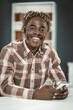 © Svyatoslav Lypynskyy - Handsome Man with Dreadlocks on His Head and in a Brown Plaid Shirt Sits at a Table with his Mobile Phone in his Hands. Broad Smile. Close-up